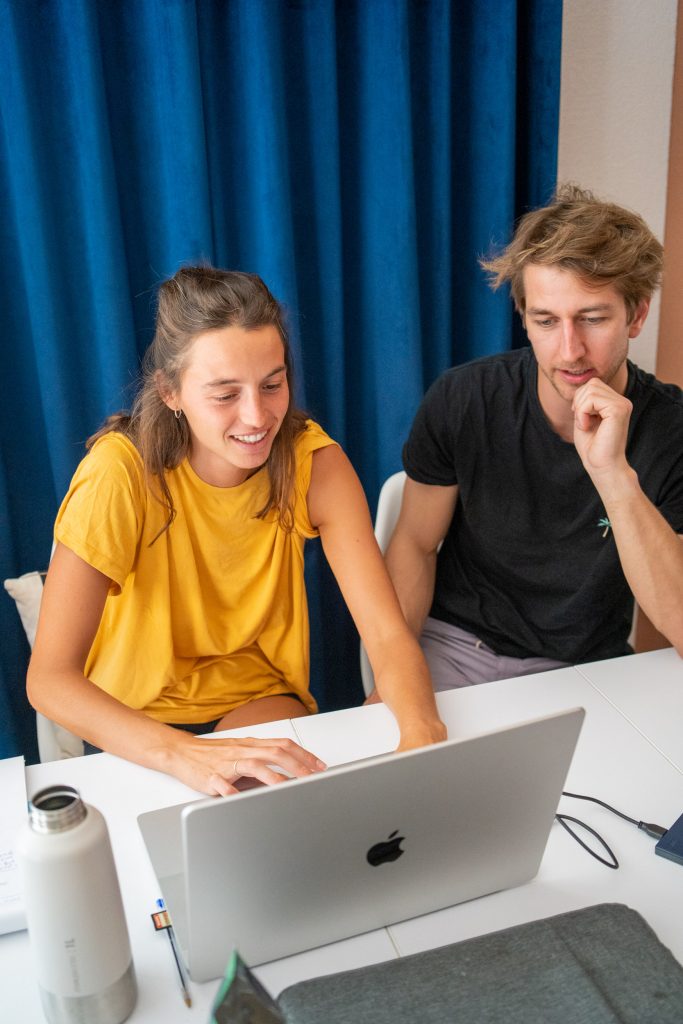 A young woman on a laptop, a young man sitting next to her, both facing the laptop's screen.