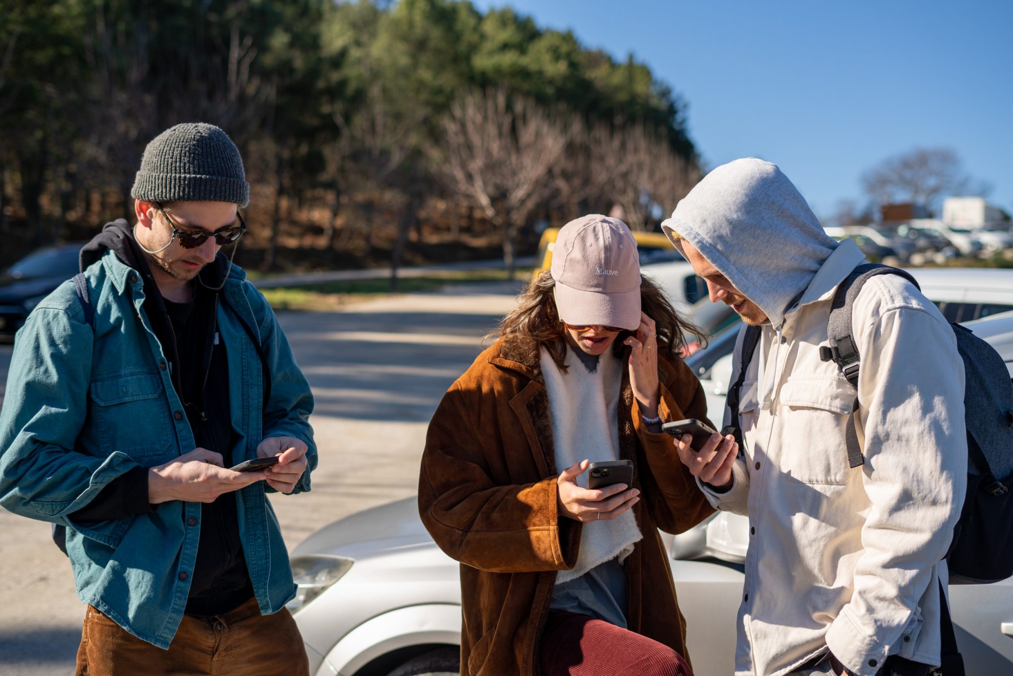 Three young people looking on their smartphones in the sun while standing in a carpark.