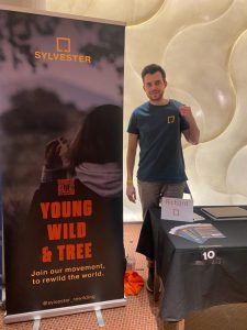 A young man with a petrol shirt smiling into the camera - next to him a roll-up with the slogan "Young Wild & Tree".