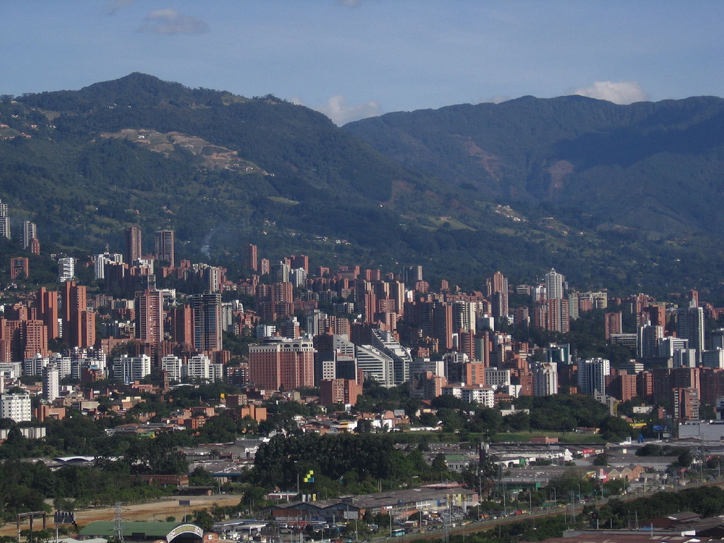 picture showing the city medellin viewed from a hill. you can see many high buildings and a mountain in the background with lots of trees